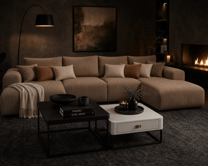 A modern sectional sofa in a warm taupe corduroy fabric, styled in a cozy, dimly lit living room with a fireplace, a black floor lamp, and two coffee tables in the foreground.