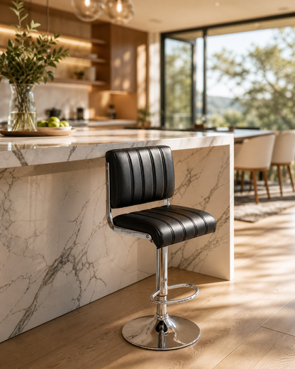 A modern black faux-leather bar stool with a chrome pedestal base and footrest, positioned in a contemporary kitchen with a marble island and large windows.