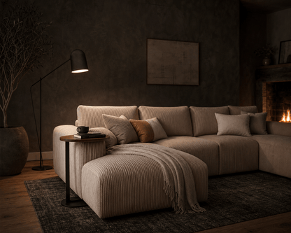A modern sectional sofa in a warm beige ribbed fabric, styled with matching throw pillows and a fringed throw blanket in a cozy, dimly lit living room with a fireplace and floor lamp.