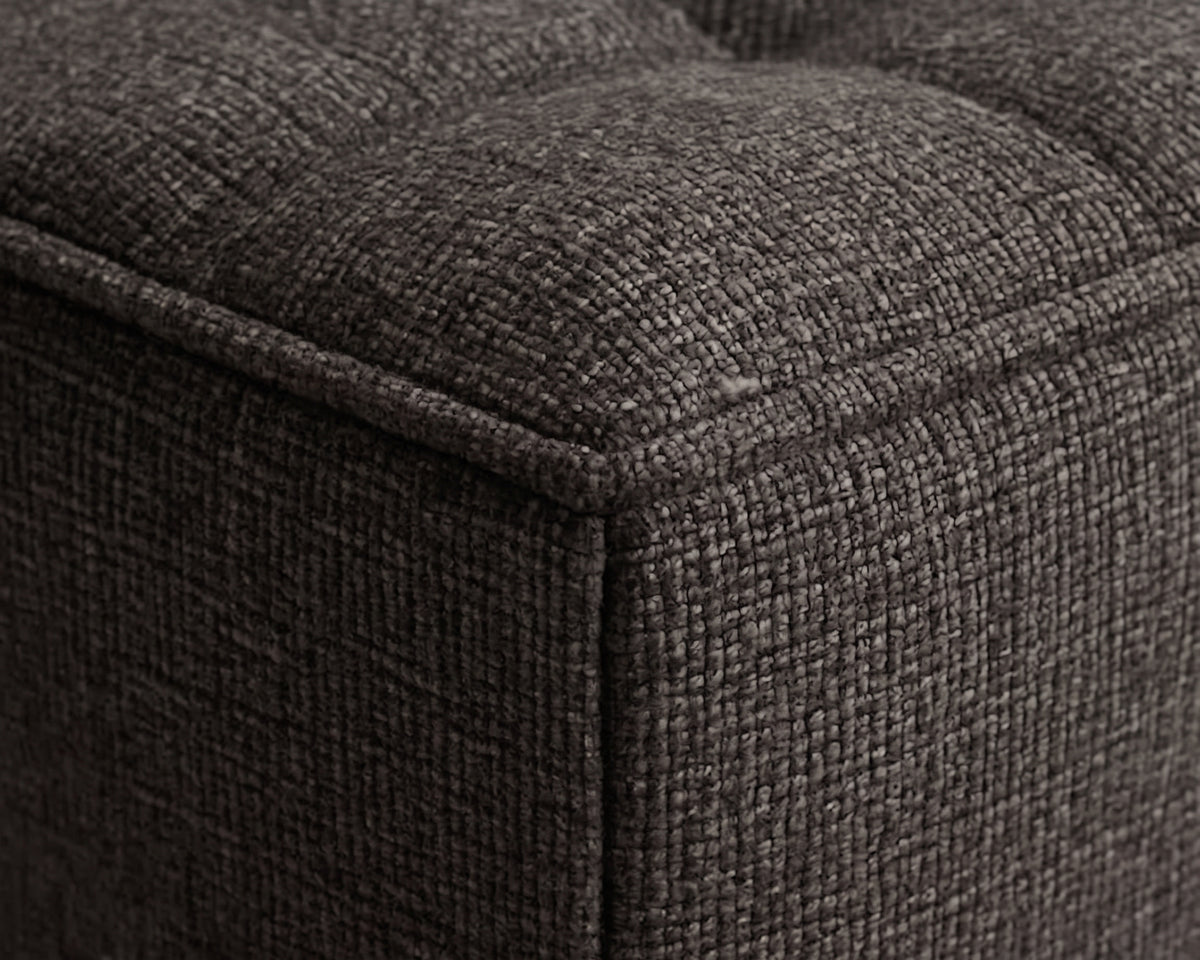 Close-up of a dark charcoal gray tufted fabric ottoman showing the textured woven upholstery and stitched corner seam.