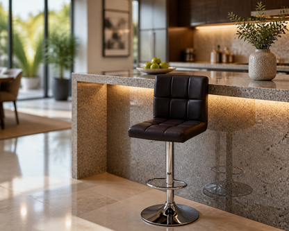A modern black bar stool with a tufted square backrest and chrome pedestal base, positioned in front of a speckled stone kitchen island in a bright, contemporary interior.