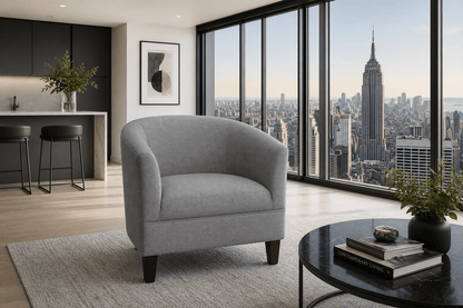 A modern gray upholstered tub chair with dark wooden legs, positioned in a high-rise apartment living room with a city skyline view of the Empire State Building through large floor-to-ceiling windows.