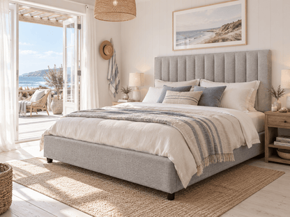 A light gray upholstered bed with a tall tufted headboard, styled with white and blue-gray bedding, a striped throw blanket, and a woven rug in a bright, modern bedroom with an ocean view.