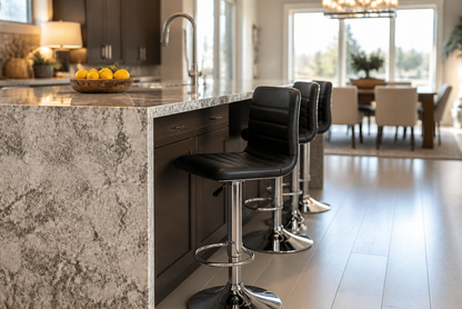 A modern kitchen island with a gray and white marbled stone countertop, dark wood cabinetry, and three black leather swivel bar stools with chrome bases, set in a bright, contemporary dining area.