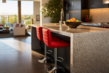 A modern kitchen island with a speckled gray stone countertop, two red bar stools with chrome bases, and a bowl of fruit on the counter, set in a bright contemporary kitchen with dark cabinetry.