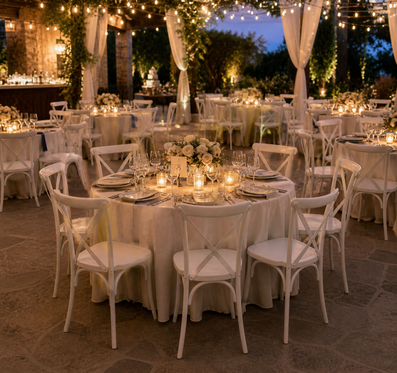 Outdoor evening event setup with round tables covered in white tablecloths, white chairs, floral centerpieces, and lit candles under warm string lights.