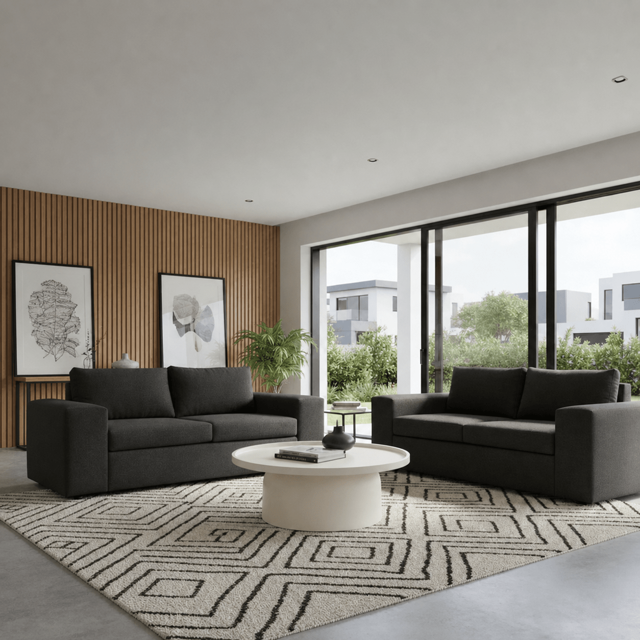 A modern living room featuring two dark gray fabric sofas arranged on a patterned rug around a round white coffee table. Large floor-to-ceiling windows with black frames look out onto greenery and buildings, and the left wall has vertical wood slats with two framed artworks.