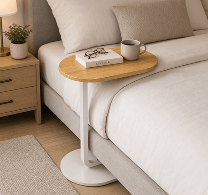A modern oval bedside table with a light wood top and white curved metal base, styled next to a bed with white linens, a mug, and a book in a bedroom setting.