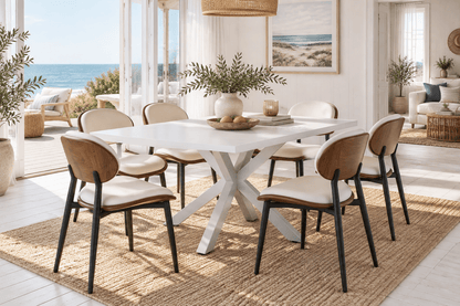 A modern dining room set featuring a white rectangular table with an X-base and six matching chairs with light beige seats, brown wood-look backs, and black legs, set on a jute rug in a bright, sunlit room with an ocean view.