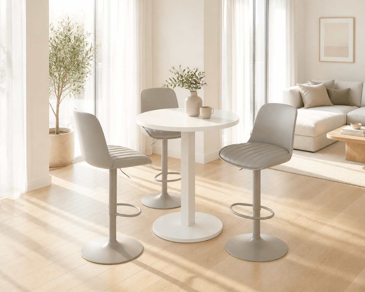 A modern dining set featuring a round white table and three light gray upholstered bar stools with metal bases, set in a bright, sunlit living room.