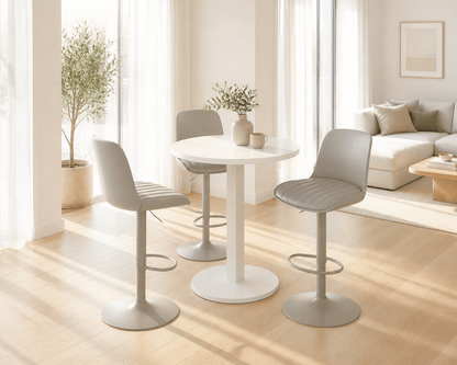 A modern dining set featuring a round white table and three light gray upholstered bar stools with metal bases, set in a bright, sunlit living room.
