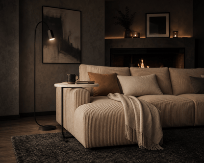A modern beige corduroy sectional sofa in a cozy living room setting with a lit fireplace, a black floor lamp, and a small side table.