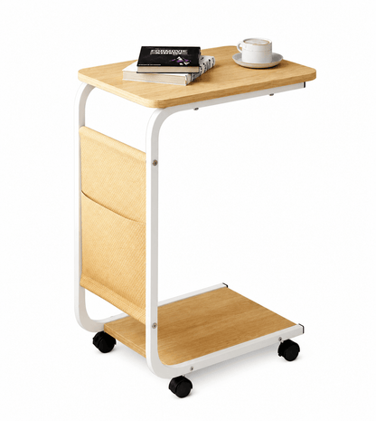 A mobile side desk with a light wood grain top, white metal frame, and black caster wheels. It features a side storage pocket, a lower shelf, and is shown with books and a coffee cup on top, set against a plain white background.
