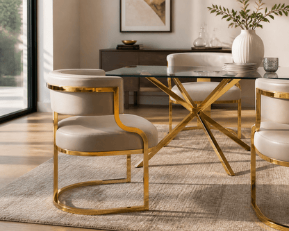 A modern dining room scene featuring a glass-top table with a gold metal X-frame base, surrounded by upholstered chairs with cream fabric and gold curved metal frames, set on a textured beige rug.