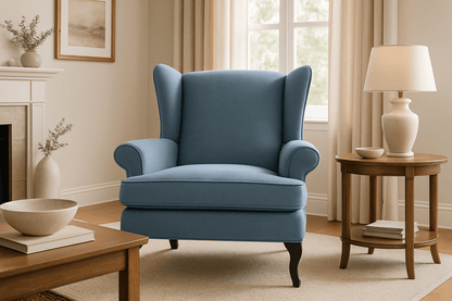 A blue wingback armchair in a beautifully styled living room with beige walls, a wooden side table, a white ceramic lamp, and a window in the background.
